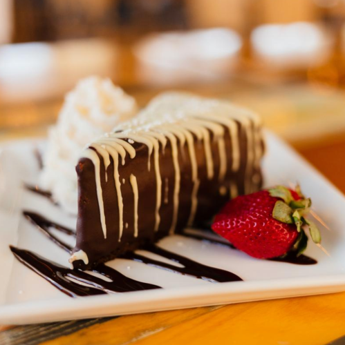 A slice of chocolate cake drizzled with white chocolate, served with a strawberry and whipped cream on a plate.